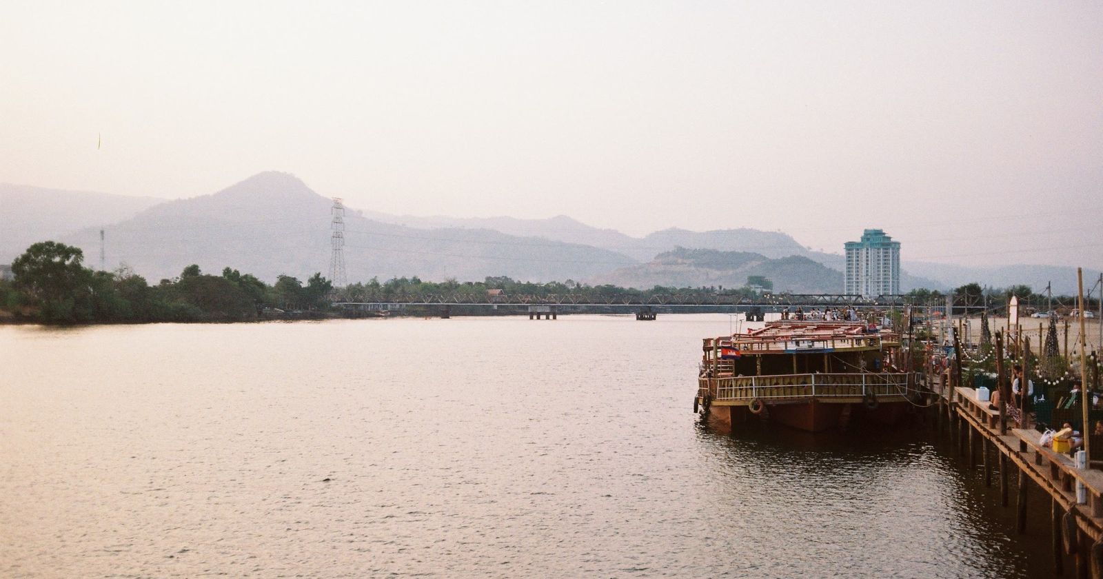 The Kampot river in soft evening light, with boats moored at a jetty and mountains behind